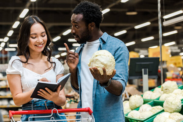 african american man gesturing and holding cabbage near asian cheerful woman with notebook 