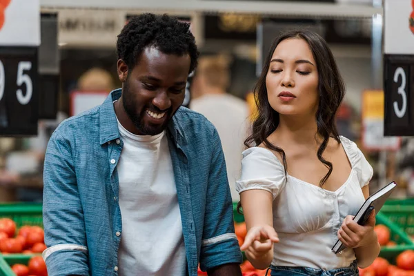 attractive asian woman with notebook pointing with finger near happy african american man