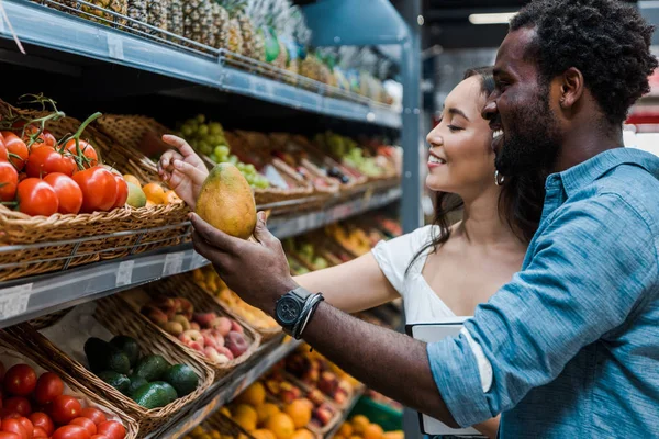 Lycklig Asiatisk Kvinna Tittar Frukter Nära Glada Afroamerikanska Mannen Butik — Stockfoto