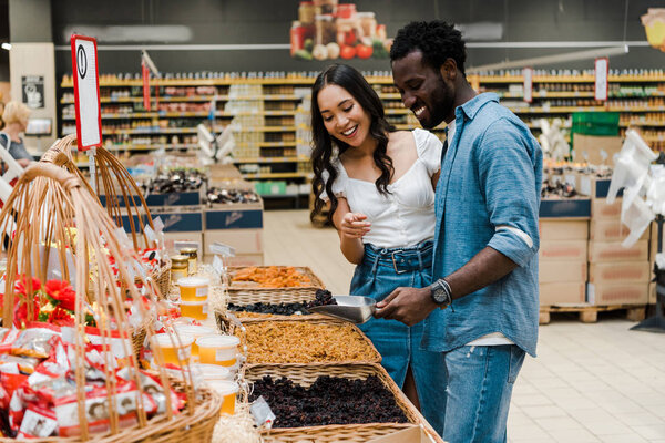happy african american man holding metal scoop with raisins near asian woman pointing with finger in store 