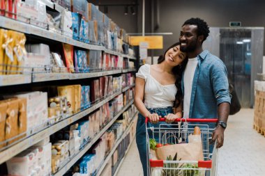 happy asian woman standing near cheerful african american boyfriend and looking at shelves with groceries   clipart