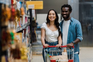 mutlu asyalı kadın ve neşeli afrikalı amerikalı adam seçici odak süpermarkette raflara bakarak 