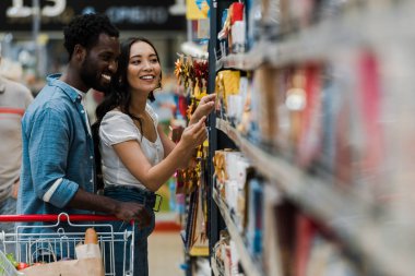 selective focus of cheerful african american man standing with asian woman pointing with finger at groceries in supermarket  clipart