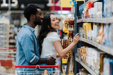 selective focus of cheerful african american man standing with cheerful asian woman pointing with finger at groceries in supermarket  clipart
