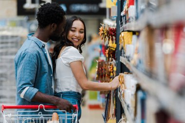 selective focus of cheerful african american man standing with cheerful asian girl smiling near groceries in supermarket  clipart