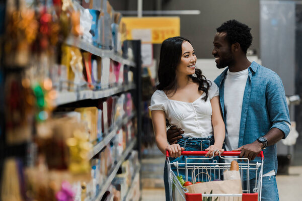 selective focus of happy asian woman looking at cheerful african american boyfriend in supermarket 