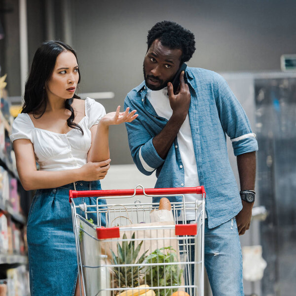 upset asian woman gesturing while looking at african american man talking on smartphone in supermarket 