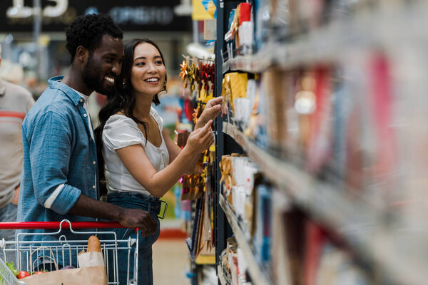 selective focus of cheerful african american man standing with asian woman pointing with finger at groceries in supermarket 