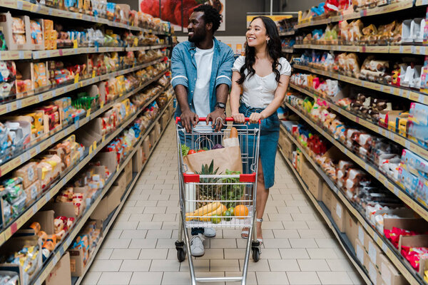 happy asian woman and african american man walking with shopping cart in supermarket 