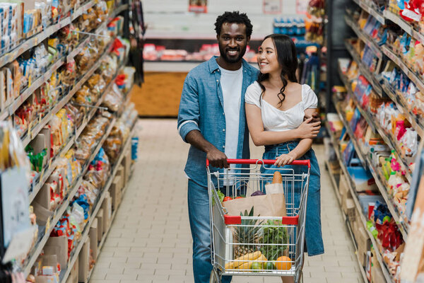 selective focus of happy asian woman and african american man walking with shopping cart in supermarket 