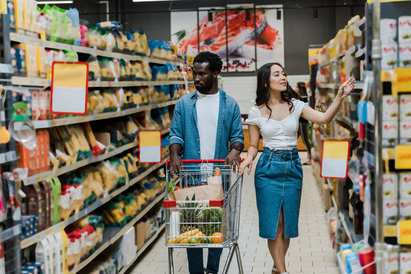 handsome man walking with shopping cart near attractive asian girl gesturing in store 