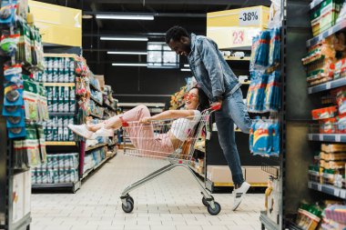 mutlu afrikalı amerikalı adam süpermarkette alışveriş sepetinde oturan asyalı kız bakıyor 