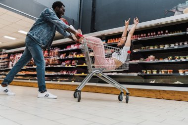 alışveriş sepetinde otururken güneş gözlüğü gesturing mutlu asya kız yakın heyecanlı afrikalı amerikalı adam düşük açı görünümü 