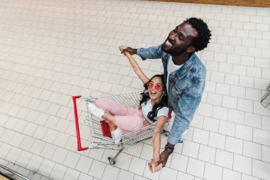 alışveriş sepeti oturan ve yakışıklı afro-amerikan adam ile el ele güneş gözlüğü neşeli asyalı kız havai görünümü 
