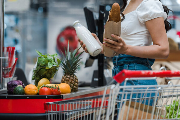 cropped view of woman holding baguette and glass bottle with milk near supermarket counter 