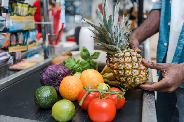 cropped view of african american man holding pineapple near supermarket counter with groceries
