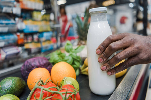 cropped view of african american man holding glass bottle with milk near supermarket counter 