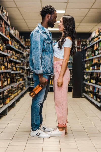 happy african american man holding bottle and looking at asian girl in supermarket 