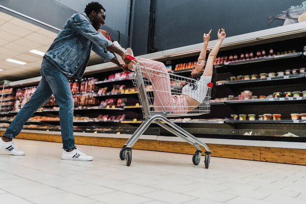 low angle view of excited african american man near happy asian girl in sunglasses gesturing while sitting in shopping cart 