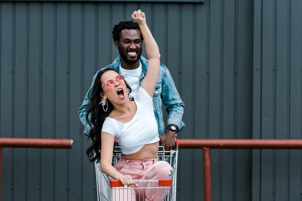 Emotional Asian Girl Gesturing While Sitting Shopping Trolley African American — Stock Photo, Image