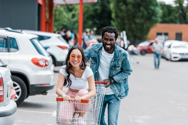 快乐的非洲美国男子走 而开朗的亚洲女孩在太阳镜坐在购物车 — 图库照片