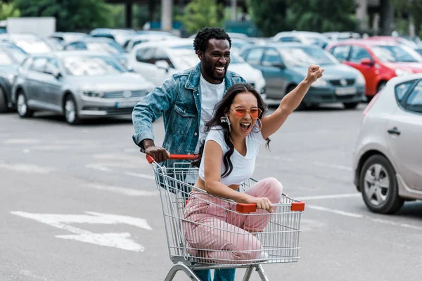 Elegante Asiático Menina Gesturing Enquanto Sentado Shopping Trolley Perto Africano — Fotografia de Stock