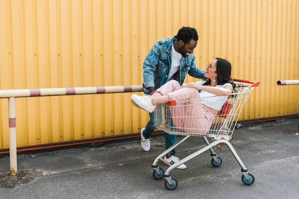 happy african american man smiling while looking at asian girl sitting in shopping trolley 