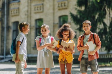 four adorable multicultural schoolchildren standing in schoolyard and holding books clipart