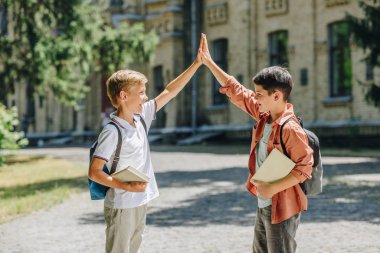 two cheerful schoolboys giving high five while standing in schoolyard clipart