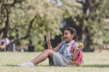 parkta çim üzerinde otururken elma tutan ve el sallayarak neşeli kız öğrenci