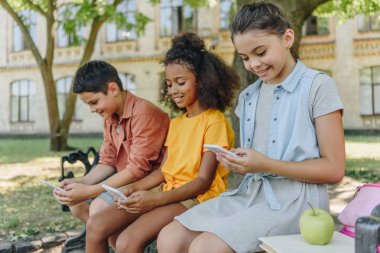 three cute multicultural schoolkids using smartphones while sitting on bench in schoolyard clipart