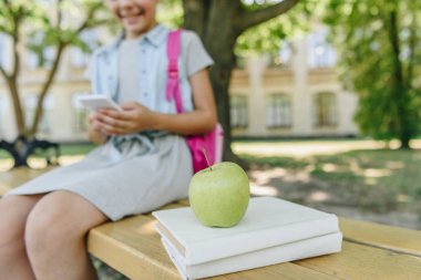 cropped view of smiling schoolgirl using smartphone while sitting on bench near books and apple clipart