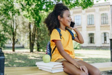 kitap ve elma yakınında bankta otururken akıllı telefon konuşurken sevimli afro-amerikan kız öğrenci