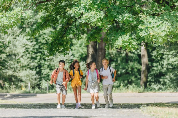 日当たりの良い公園で実行されているバックパックを持つ4人の幸せな多文化の学校の生徒 — ストック写真