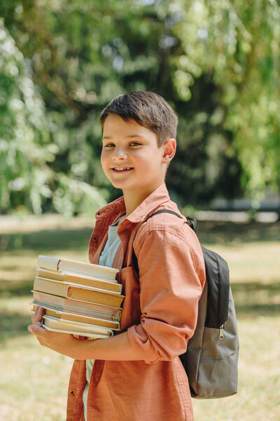 happy schoolboy smiling at camera while holding books in park