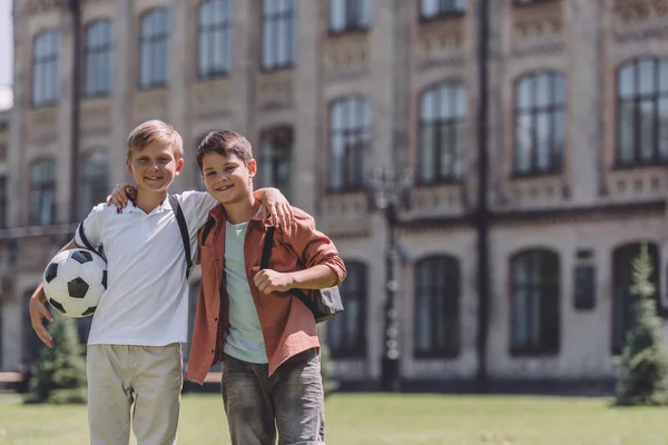 サッカーボールで学校の近くに立っている間に抱きしめる2人の陽気な男子生徒 — ストック写真