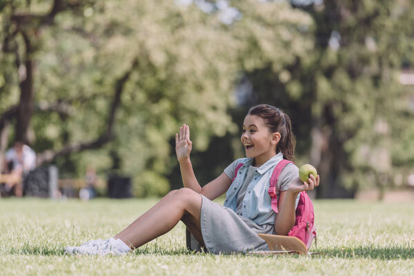 cheerful schoolgirl holding apple and waving hand while sitting on lawn in park