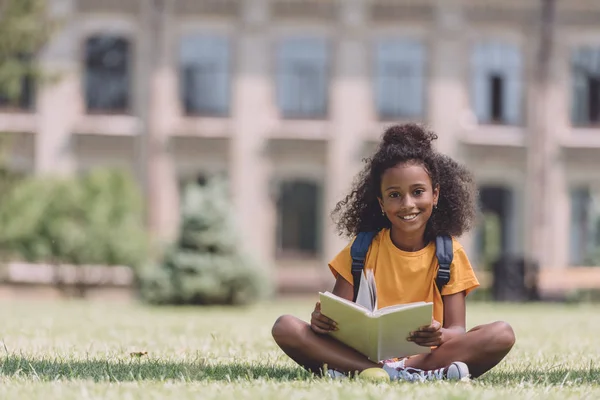 本と芝生の上に座っている間 カメラで微笑むかわいいアフリカ系アメリカ人の女子高生 — ストック写真