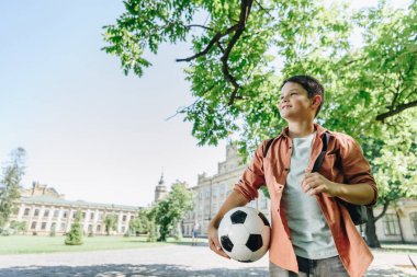 arka planda mavi gökyüzü ve yeşil ağaç ile parkta yürüyen futbol topu ile sevimli okul çocuğu