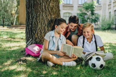 üç sevimli sınıf arkadaşları kitap okuma ve ağacın altında çim üzerinde otururken gülümseyerek