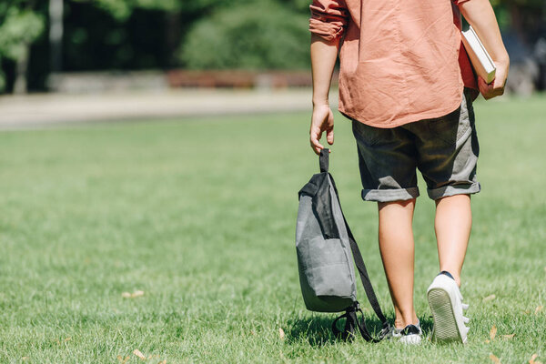 cropped view of schoolboy with backpack walking on lawn in park