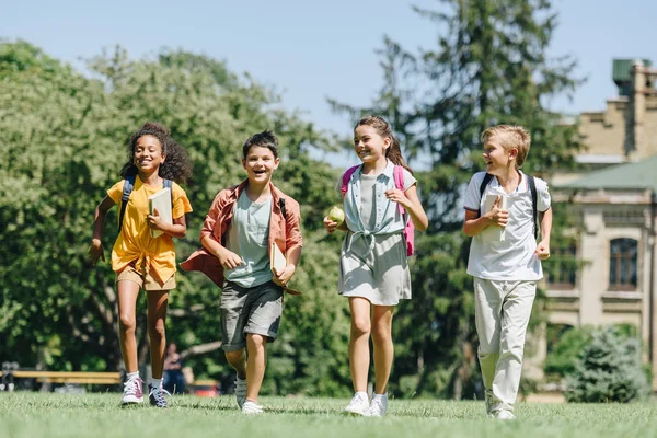 公園の芝生の上を一緒に走る4人の幸せな多文化の小学生 — ストック写真