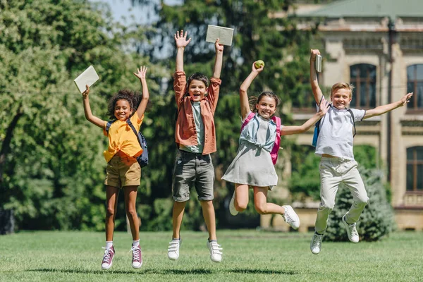 四个兴奋的多民族学童在公园草坪上拿着书时跳 — 图库照片