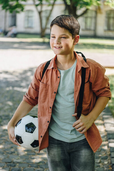 cheerful schoolboy looking away while walking in park and holding soccer ball
