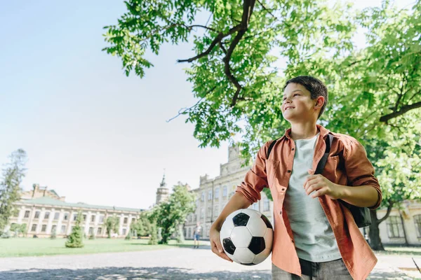 青い空と緑の木を背景に公園を歩くサッカーボールを持つかわいい男子生徒 — ストック写真