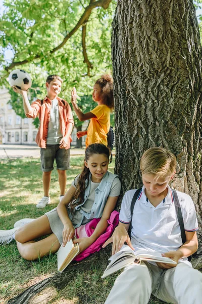 Bedårande Skolbarn Sitter Underträd Och Läsa Böcker Nära Mångkulturella Vänner — Stockfoto