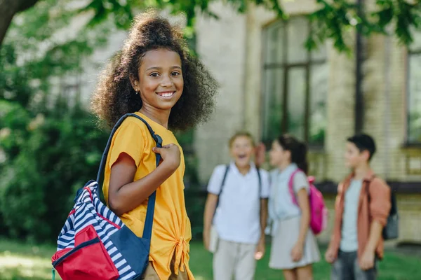 多民族の友人の近くのカメラで笑顔のバックパックと陽気なアフリカ系アメリカ人の女子高生の選択的な焦点 — ストック写真
