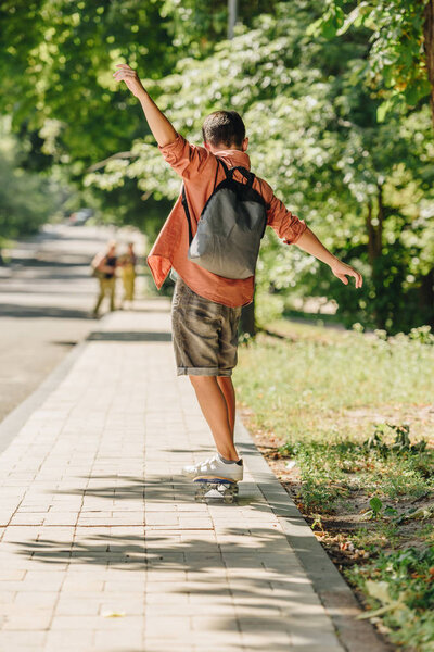 back view of schoolboy with backpack riding skateboard on sunny street