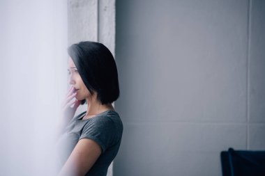 beautiful depressed woman looking through window at home with copy space clipart