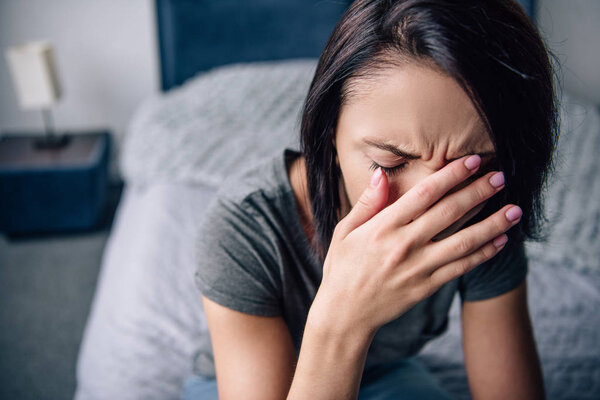 depressed woman sitting on bed at home, crying and covering face with hands